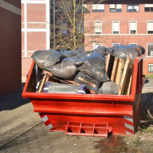 Full orange metal Skip container standing on the road near the house. In the container are old things, suitcases, furniture, black plastic bags of garbage and rubbish after cleaning the house.